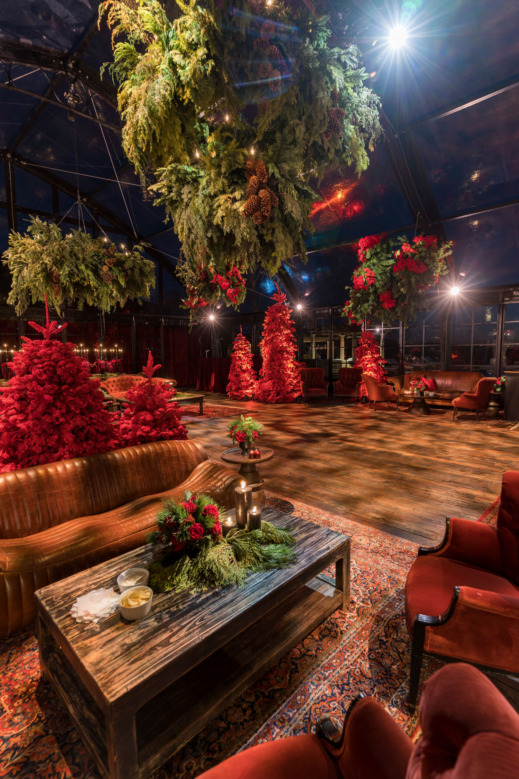 Side view of modern cognac leather tufted sofa next to low wood coffee table and red velvet chairs on vintage Persian rugs surrounded by ferns and poinsettia in a glass tent at Kourtney Kardashian Barker&#8217;s 2022 Christmas Eve party Kourtney kardashian barker&#8217;s 2022 xmas eve party glass tent wood flooring and gorgeous lush plants hanging from ceiling